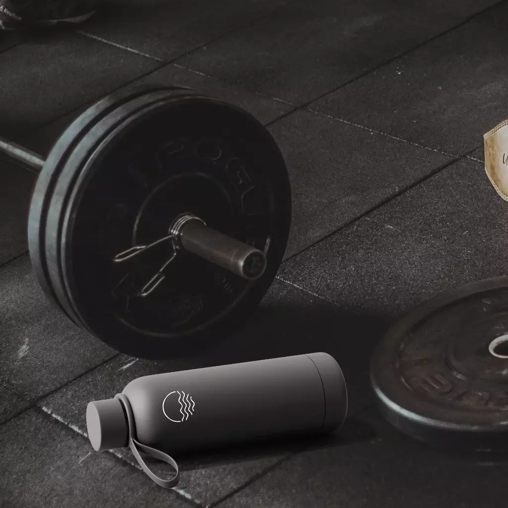“Graphite black HydroTrak smart water bottle placed on a gym floor beside weightlifting equipment.”